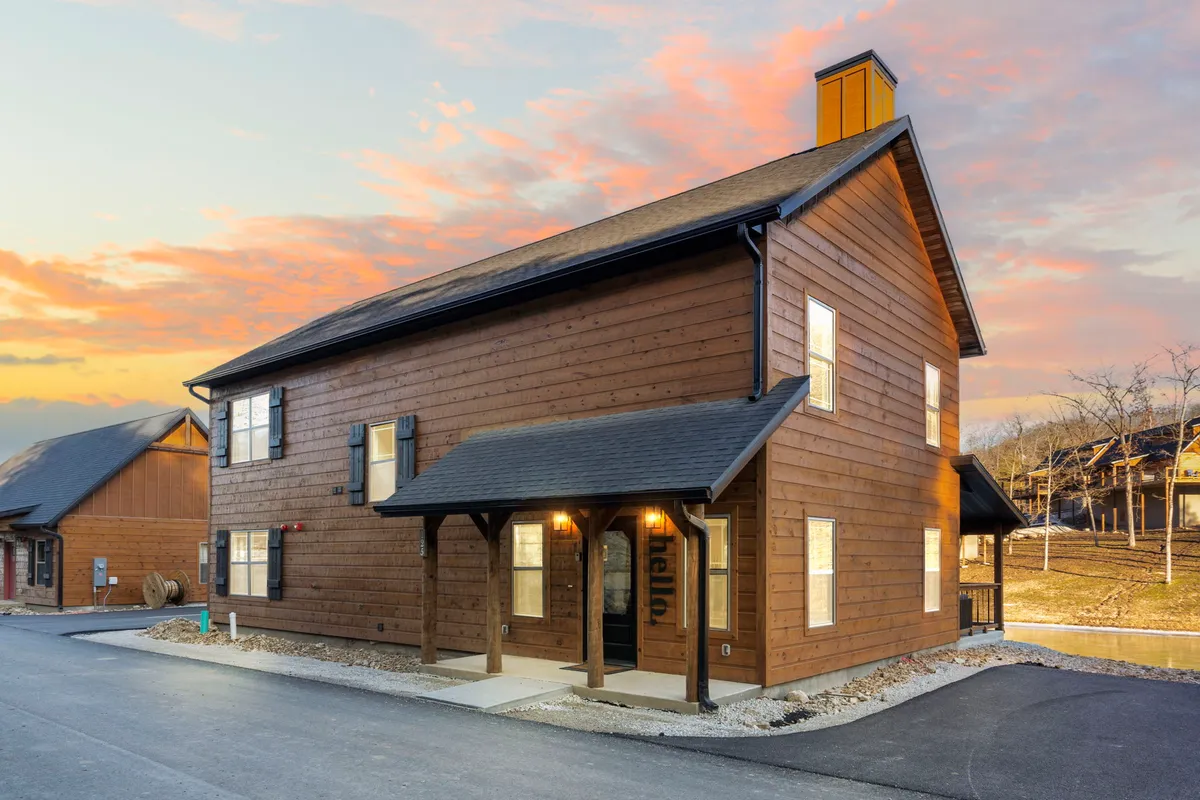 A brand-new wooden cabin exterior is shown at sunset, with warm lighting glowing from within and inviting front porch seating. The background features natural surroundings of trees and open sky, capturing an atmosphere of relaxation and welcome.