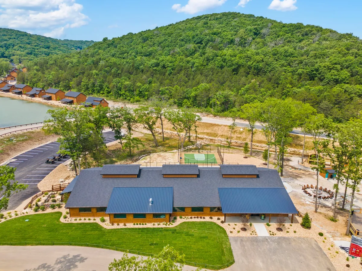 An exterior view featuring the main building with a dark roof, lush greenery, and a clearly visible recreational area with a sports court. Families are seen engaging in outdoor activities, suggesting energetic play and group enjoyment.
