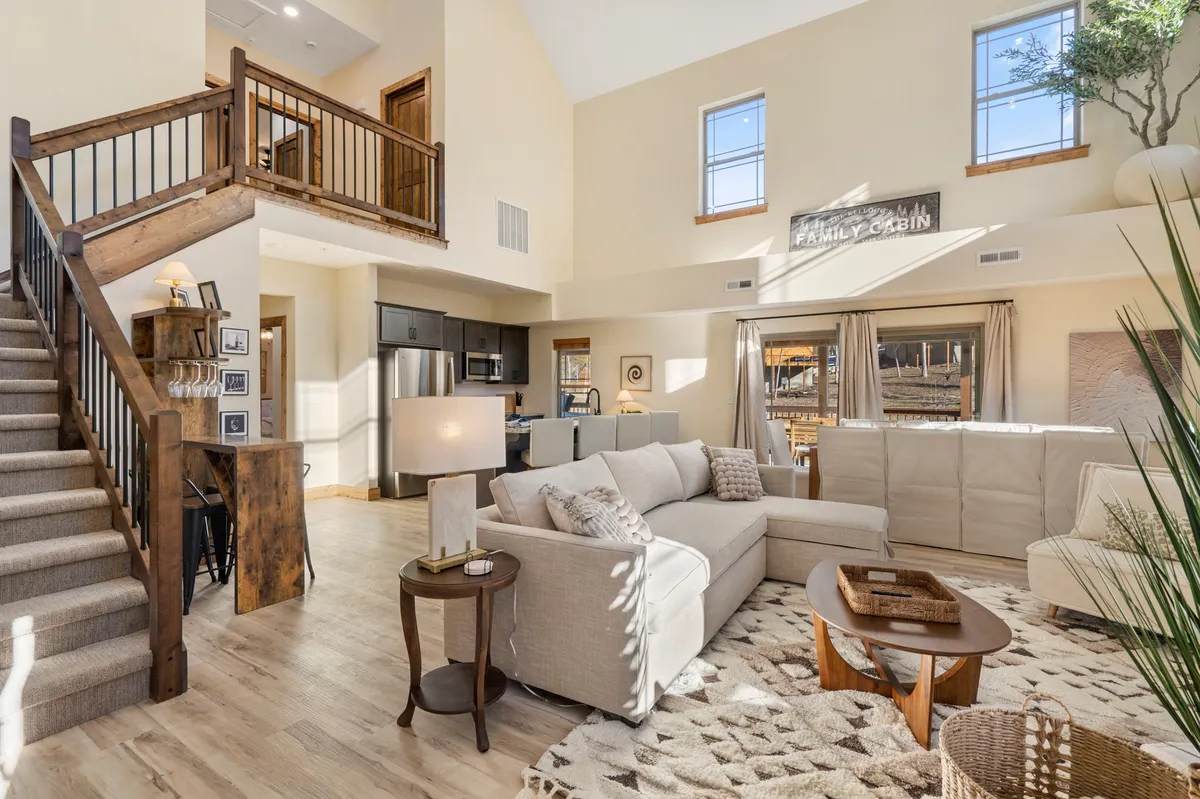 The image features a spacious, warmly lit living area with a large sectional sofa perfect for gatherings, a rustic wooden dining table for shared meals, a modern kitchen, and farmhouse-inspired decor—all set beneath exposed wood beams and next to a staircase leading to more private spaces.