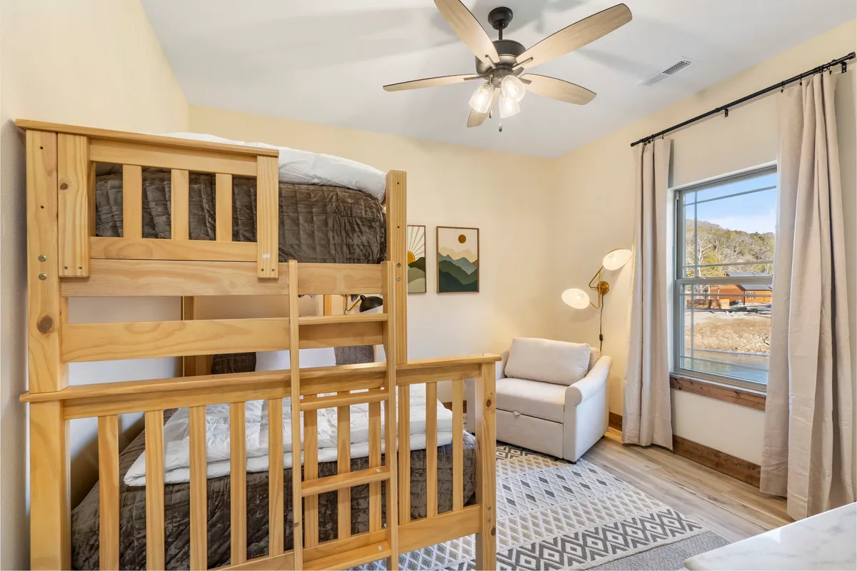 A cheerful bunk room featuring a sturdy bunk bed, a cozy armchair, window view, and bright decorative art. The space is inviting and designed for children's comfort and play.