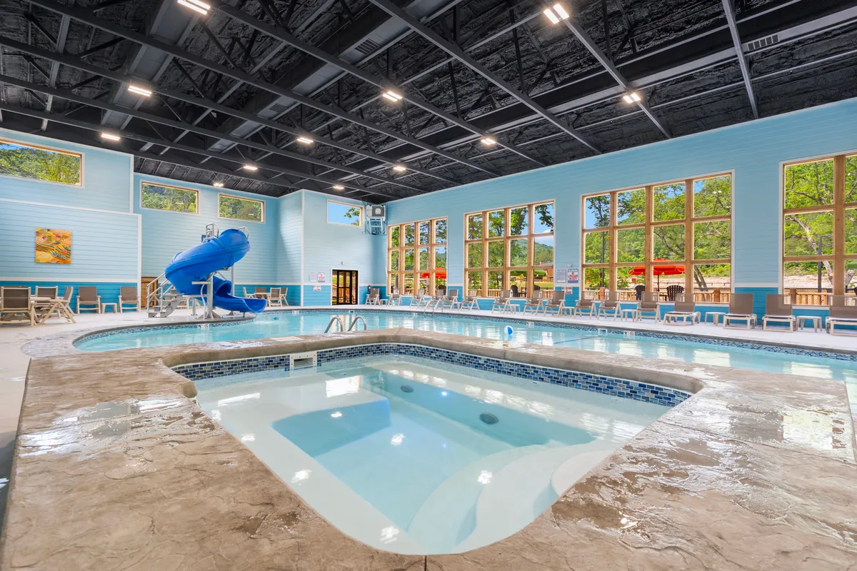 A bright, spacious indoor pool area featuring a water slide, jacuzzi, large windows, and ample poolside seating. Families and children are seen swimming, playing, and relaxing, creating a joyful and lively atmosphere.