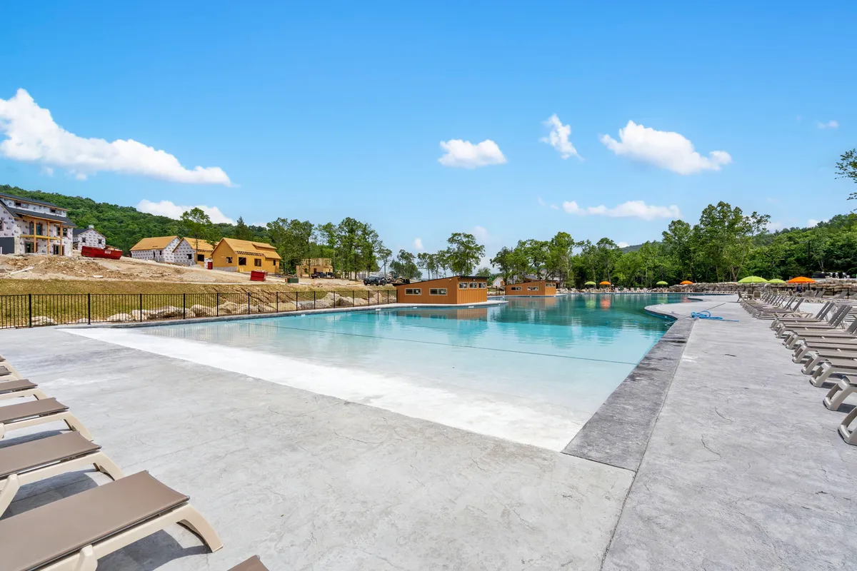 A large outdoor swimming pool surrounded by loungers, colorful umbrellas, and lush trees. The area is bustling with people swimming, sunbathing, and socializing, exuding a leisurely and communal summer vibe.