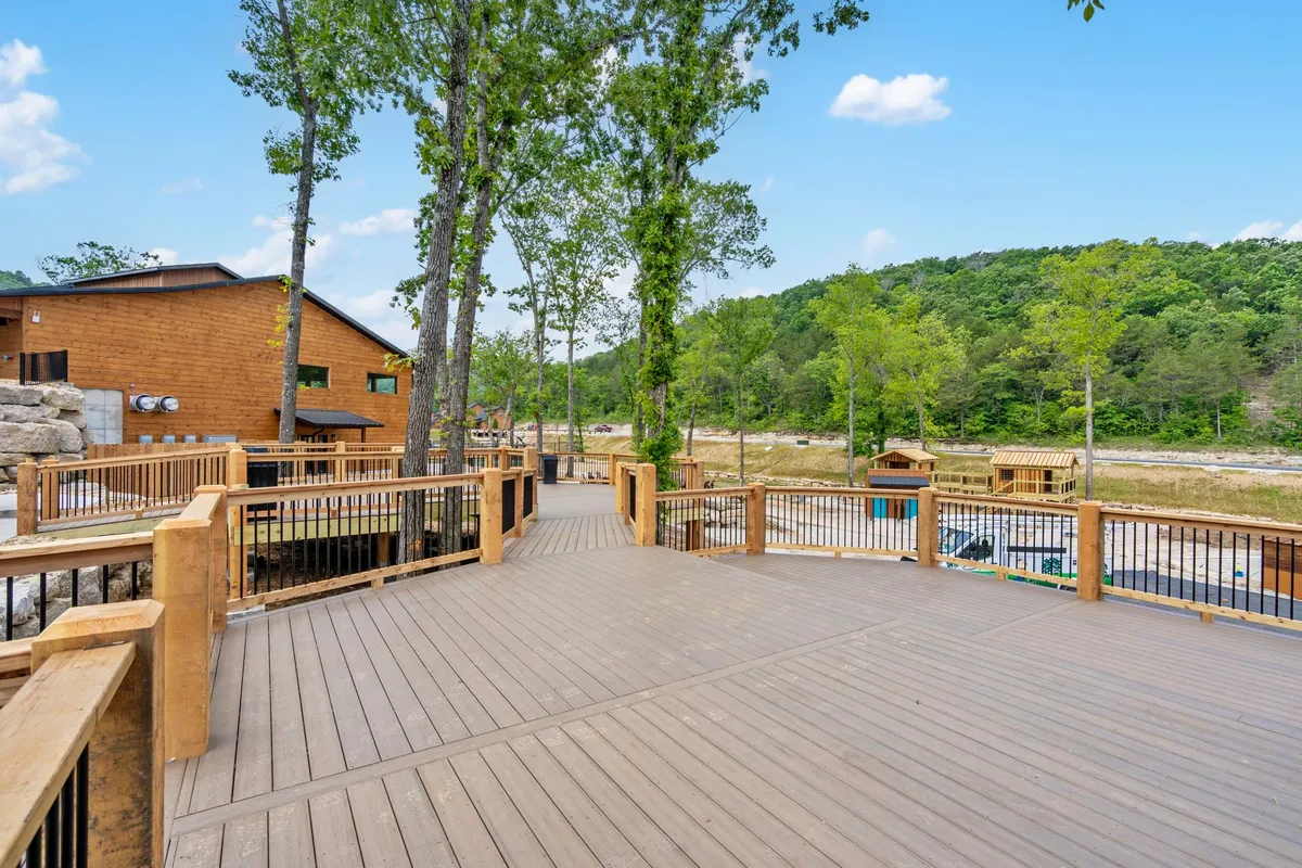 This image showcases a wooden deck with a clear view of a gentle river below, framed by tall trees and natural foliage. The spacious deck is perfect for gathering, outdoor meals, or simply unwinding to the sound of flowing water.