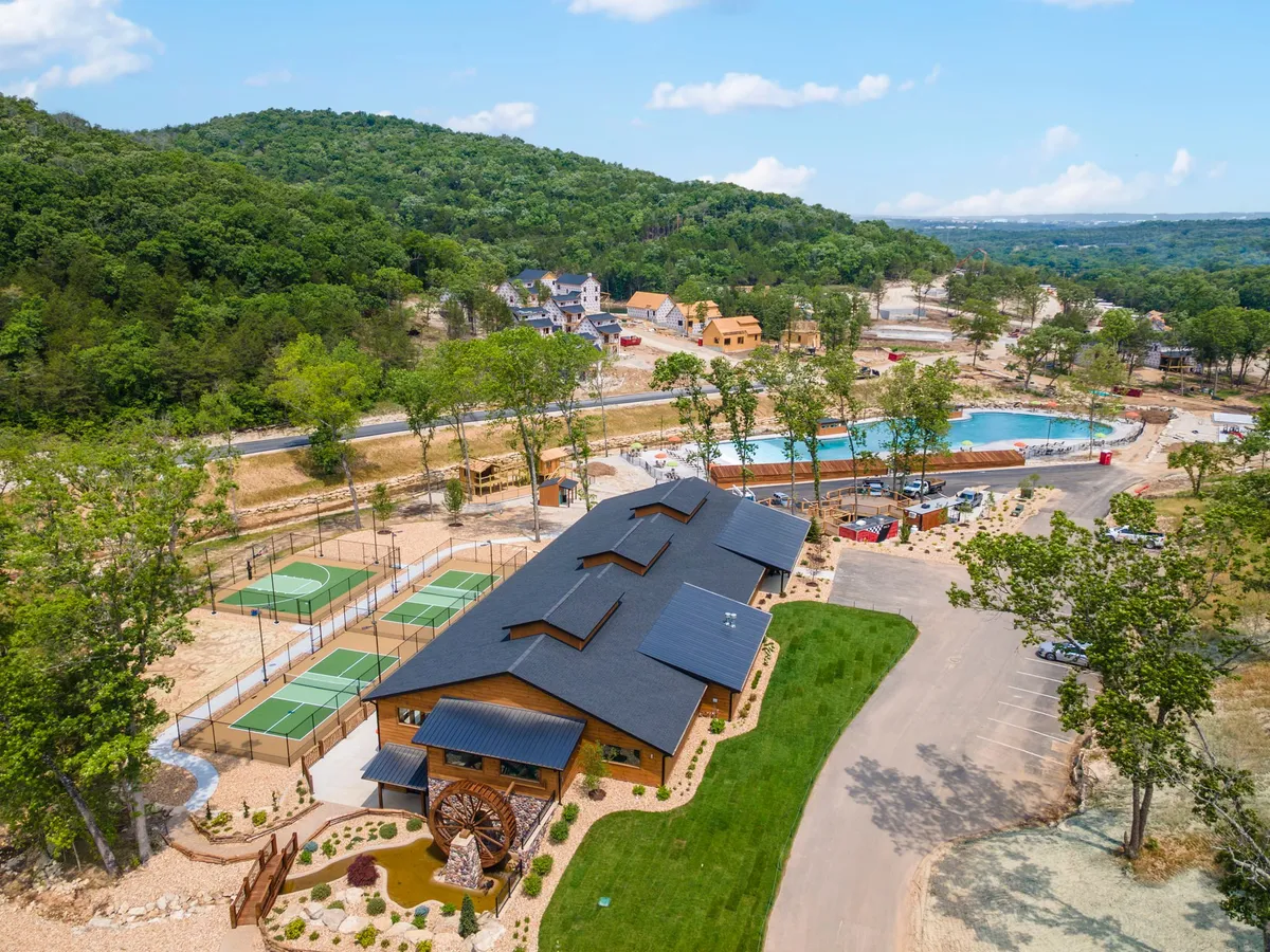 An expansive view of the resort community shows a large swimming pool, tennis courts, and the main lodge, all nestled among rolling green hills. The area feels open and recreational, highlighting communal amenities and outdoor activities.