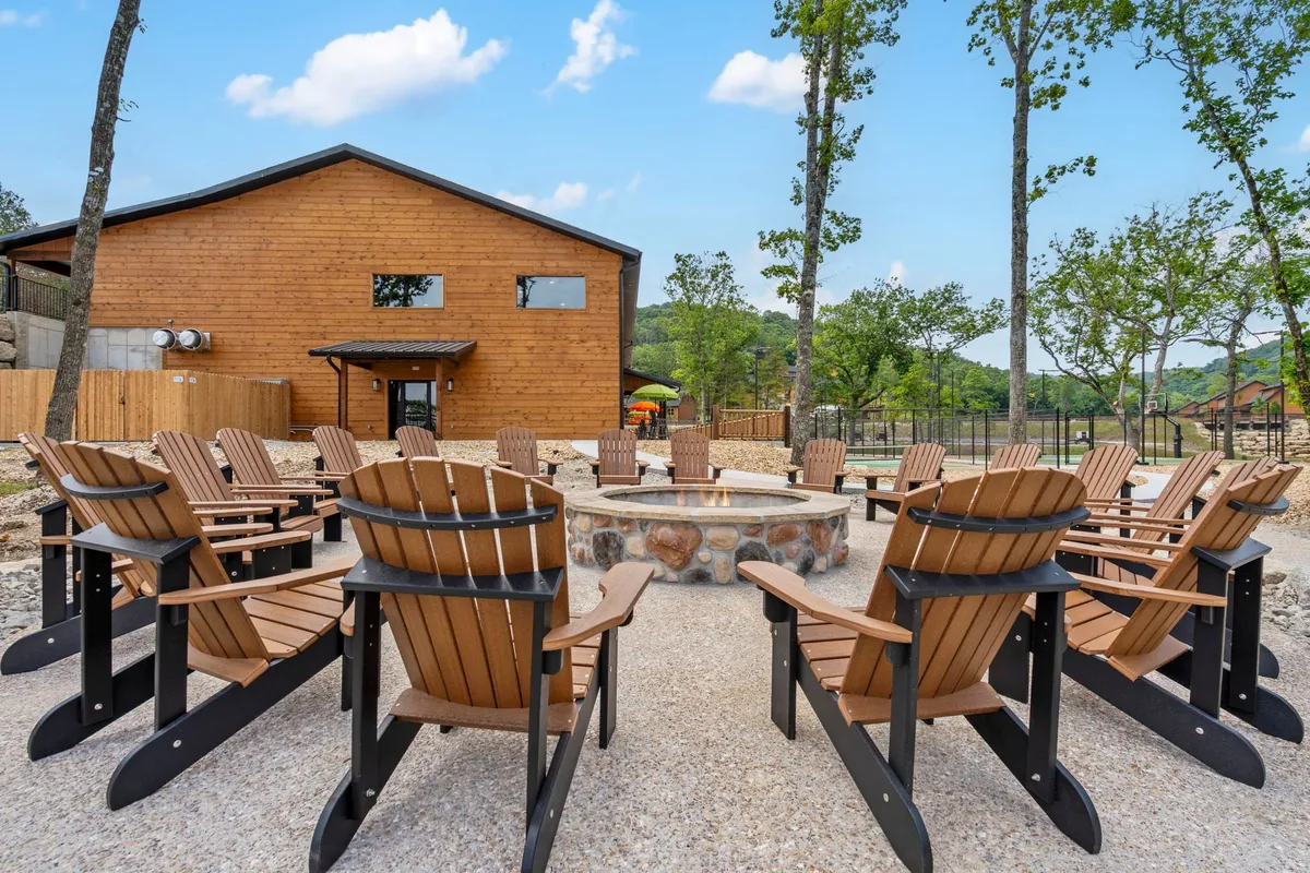 A cozy outdoor fire pit area with Adirondack chairs surrounding a crackling fire, nestled beside a classic wooden cabin. The background features trees and open sky, creating a relaxed and inviting atmosphere ideal for group gatherings in the evening.