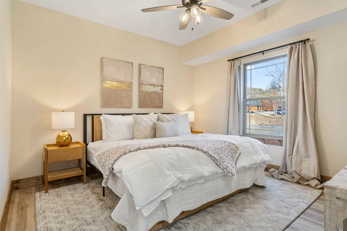 A serene king-sized bedroom with plush bedding, decorative paintings above the headboard, a nightstand with a lamp, and a window letting in natural light. The setting feels private, comfortable, and perfect for unwinding after a long day.