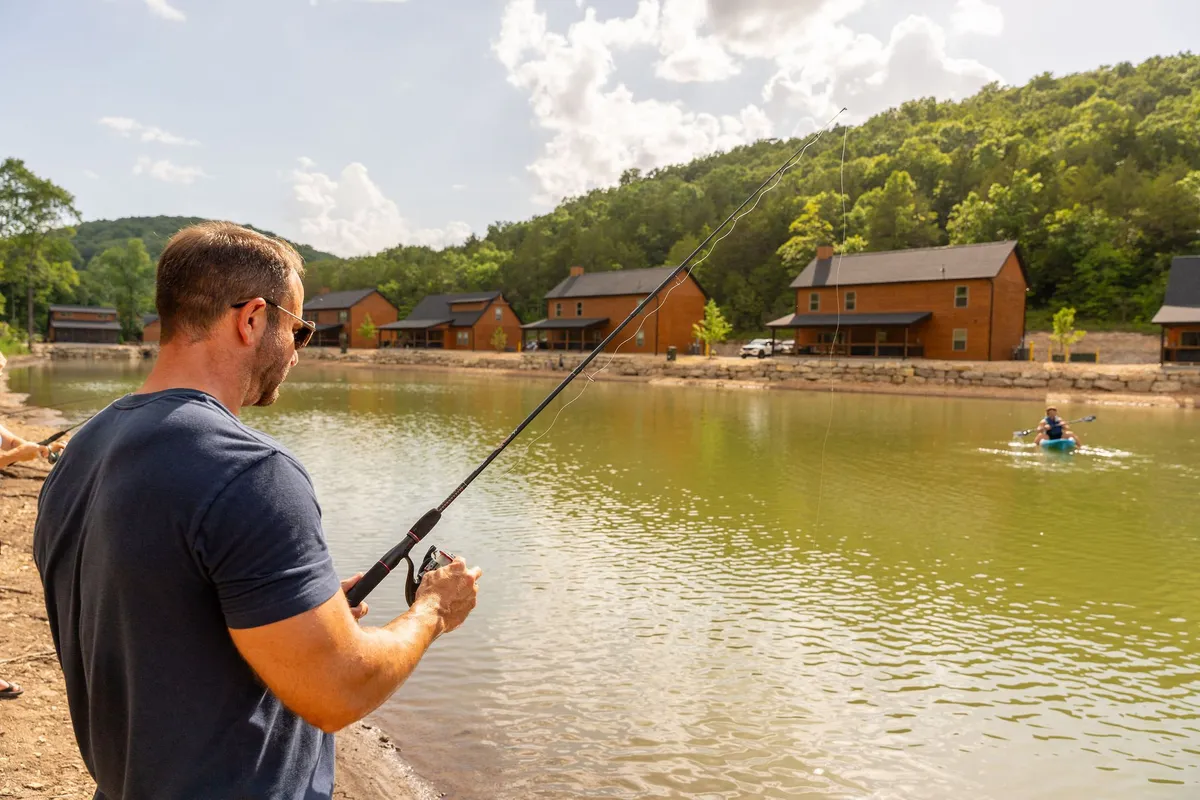 A peaceful lakeside setting where a man is fishing by calm water, while another person enjoys kayaking. Wooden cabins are tucked beside the shoreline, and soft light bathes the landscape. The gentle activity on the water and the rustic cabins create an inviting atmosphere for outdoor recreation and relaxation.